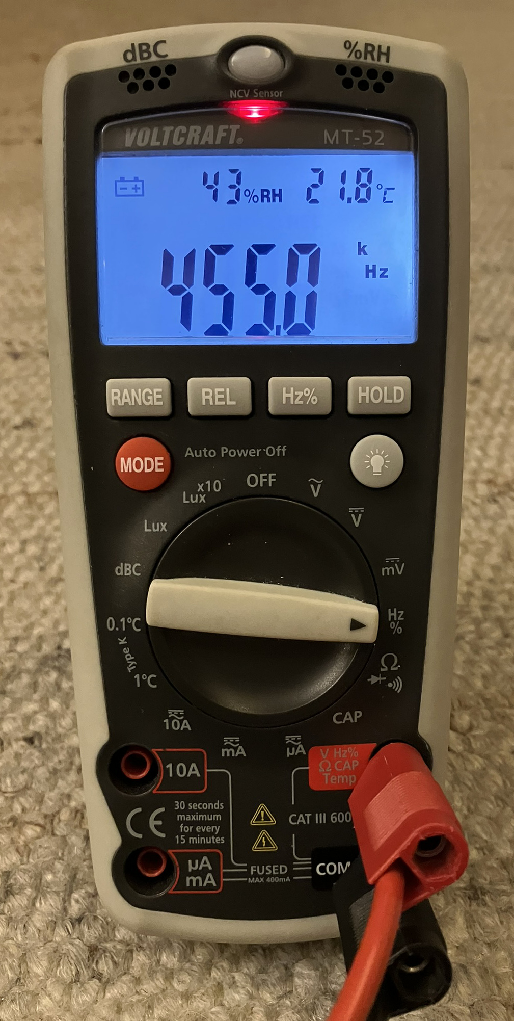 Dieser Alt-Text wurde noch nicht überprüft.
<p>Kurzfassung: Ein tragbares Digitalmultimeter mit blau beleuchtetem Display zeigt 455.0 kHz sowie 43 % relative Luftfeuchte und 21.8 °C an.</p>
<p>Detaillierte Beschreibung: Das Gerät im grauschwarzen Gummischutzgehäuse liegt auf einem beigen Teppich. Oben leuchtet eine rote LED neben der Aufschrift „NCV Sensor“; darüber sind die Hinweise „dBC“ und „%RH“ mit kleinen Lüftungsöffnungen zu sehen. Auf dem Display steht links ein Batteriesymbol, daneben „43%RH“ und rechts „21.8°C“; die großen Ziffern in der Mitte zeigen „455.0“, flankiert von den Einheiten „k“ und „Hz“. Unter dem Display befinden sich Tasten mit den Beschriftungen „RANGE“, „REL“, „Hz%“ und „HOLD“, links eine rote „MODE“-Taste und rechts eine Taste mit Lampensymbol. Der zentrale Drehschalter steht auf der rechten Seite in der Position mit der Beschriftung „Hz %“; weitere Skalen rundherum sind u. a. mit „V“, „mV“, „CAP“ und „Lux“ markiert. Unten sind vier Buchsen zu sehen: „10A“, „µA mA“, „COM“ und „V Hz Ω CAP Temp“; in „COM“ steckt ein schwarzer Stecker, in der rechten Buchse ein roter Stecker mit Kabel, daneben Sicherheits- und Messkategorienhinweise wie „CAT III 600V“ und „FUSED MAX 400mA“. Auf der Front sind außerdem die Markierungen „VOLTCRAFT“ und „MT-52“ sowie „Auto Power Off“ sichtbar.">
<figcaption>Abbildung NE-13.13.1: Multimeter, das im Frequenzmessbereich $455 kHz$ anzeigt. Darüber erscheinen ein Symbol für niedrige Batteriespannung, die Luftfeuchtigkeit und die Temperatur. Diese Werte haben nichts mit der Frequenzmessung zu tun.</figcaption></p>
</figure>
</div>
</section>
<section></section>
<section><ul>
<li>Anzeige der Frequenz</li>
<li>Bei älteren Geräten steht hinten ein $10^x$-Multiplikator
</left></li>
</ul>
<div class=