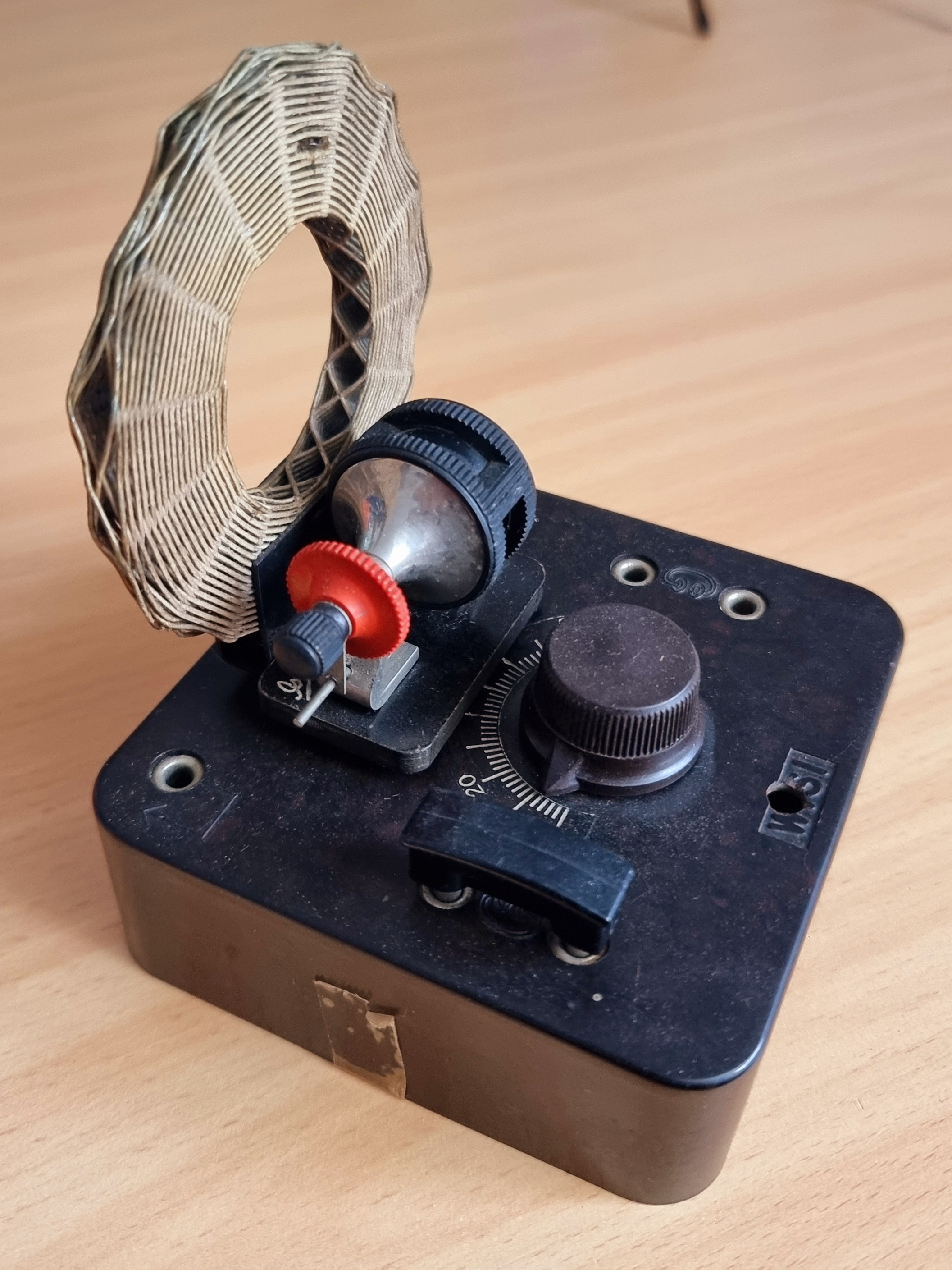 Dieser Alt-Text wurde noch nicht überprüft.
<p>Kurzfassung: Ein schwarzes, älter wirkendes Tischgerät mit senkrechter Ringspule und mehreren Einstellelementen steht auf einer hellen Holzoberfläche.</p>
<p>Detaillierte Beschreibung: Auf einem rechteckigen, dunkelbraunen/ schwarzen Gehäuse (mit abgenutzter Oberfläche) sitzt links eine flache, aufrecht stehende Ringspule aus eng gewickeltem, hellem Drahtband mit rautenförmigem Muster. Davor befinden sich ein kleiner schwarzer Einstellmechanismus mit rotem gerändelten Rädchen und ein glänzendes, halbkugeliges Metallteil. Rechts ist ein großer, gerändelter Drehknopf neben einer halbkreisförmigen Skala mit Markierungen (u. a. 0–20), darüber ein schwarzer Schieber als Zeiger. In der rechten oberen Gerätefläche liegen zwei versenkte Metallschrauben/Anschlüsse; an zwei Ecken sind Bohrungen sichtbar. An der Frontseite klebt ein kleines Stück vergilbtes Klebeband. Der Hintergrund ist unscharf, die Holzplatte zeigt feine Maserung.">
<figcaption>Abbildung E-10.1.1: Detektor-Empfänger</figcaption></p>
</figure>
</aside>
Beim Detektor-Empfänger wird ein Parallel-Schwingkreis, bestehend aus Spule und abstimmbarem Kondensator, durch ein empfangenes Rundfunksignal <a class=