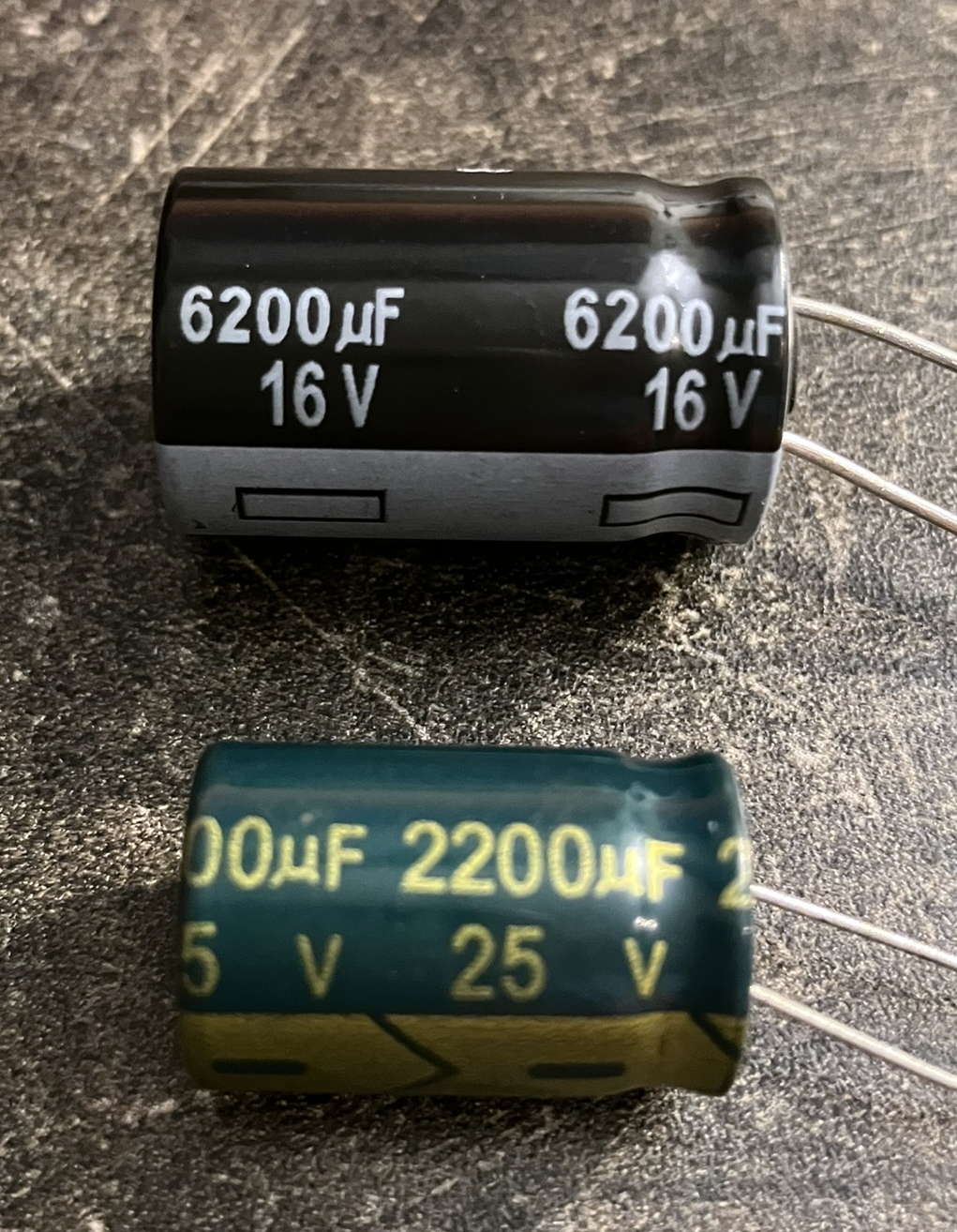 Dieser Alt-Text wurde noch nicht überprüft.
<p>Kurzfassung: Zwei liegende Elektrolytkondensatoren mit Drahtanschlüssen auf einer dunklen, rauen Arbeitsfläche.</p>
<p>Detailbeschreibung: Oben ein größerer, zylindrischer Kondensator mit schwarzem Gehäuse und grauem Ring am unteren Ende; in weißer Schrift steht mehrfach „6200 µF 16 V“, die beiden Anschlussdrähte ragen nach rechts heraus. Darunter ein etwas kleinerer, dunkelgrüner Kondensator mit gelb-goldener Aufschrift „2200 µF 25 V“ und zusätzlichen Markierungen auf einem olivfarbenen Streifen; auch hier führen zwei Drähte nach rechts. Die Oberfläche zeigt Kratzer und Staub, das Licht erzeugt glänzende Reflexe auf den Kondensatoren, die nahezu parallel zueinander liegen.">
<figcaption>Abbildung NE-6.11.2: Elektrolytkondensatoren mit den Spannungsfestigkeiten 16 Volt und 25 Volt</figcaption></p>
</figure>
</aside>
Eine weitere Kenngröße ist der *Spitze-Spitze-Wert*. Das ist der Unterschied zwischen dem höchsten und dem niedrigsten Ausschlag. Für Sinusförmige Wechselspannungen gilt:
<p>$U_\text{SS} = 2\cdot \hat{U}$.</p>
<section class=