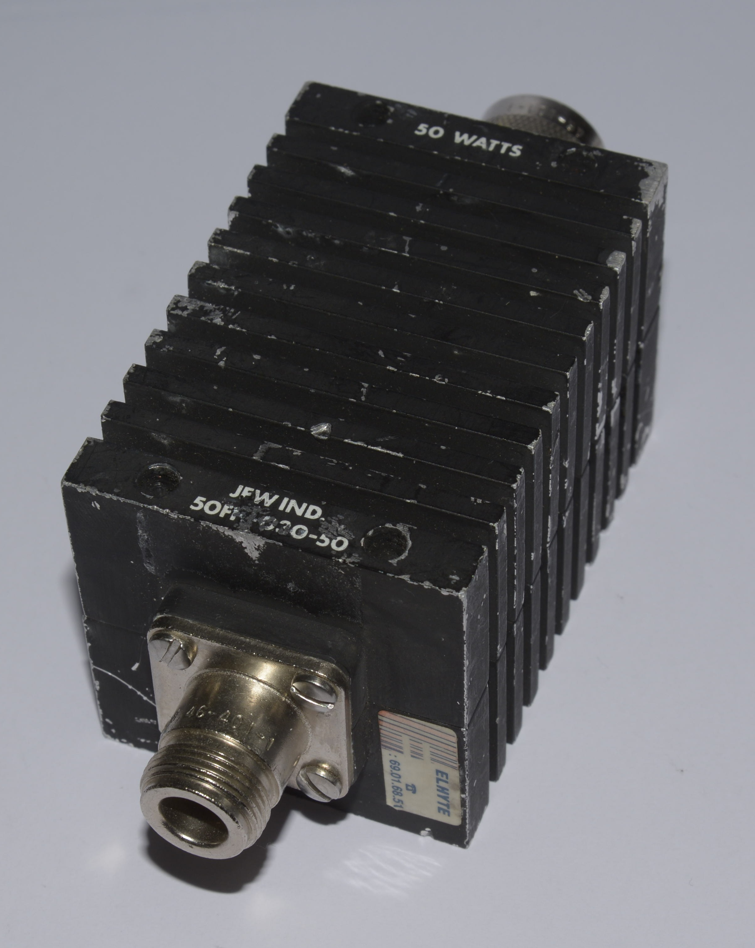 Dieser Alt-Text wurde noch nicht überprüft.
<p>Kurzfassung: Schwarzer, quaderförmiger Metallblock mit dichten Kühlrippen, der Aufschrift „50 WATTS“ und Koaxialanschlüssen an beiden Enden.</p>
<p>Detaillierte Beschreibung: Das Objekt liegt schräg auf einem hellgrauen Hintergrund und besteht aus einem schwarzen, gerippten Kühlkörper mit mehreren parallelen Längsrippen. An der Vorderseite sitzt ein koaxialer Einbausteckverbinder auf einer rechteckigen Metallflanschplatte, befestigt mit zwei Schrauben; die Lackierung ist an Kanten und Flächen sichtbar angeschlagen. Auf der Stirnseite sind die Worte „JFW IND.“ sowie eine teils abgeriebene Modellbezeichnung zu lesen. Auf der Oberseite steht „50 WATTS“. Auf der rechten Seitenfläche klebt ein kleiner Aufkleber mit der Aufschrift „ELMITE“ und einer Zahlenfolge. Hinten ist ein weiterer koaxialer Anschluss teilweise sichtbar. Das Licht fällt von oben links ein und erzeugt Schatten in den Rippenzwischenräumen.">
<figcaption>Abbildung NE-13.6.1: Ein Dämpfungsglied</figcaption></p>
</figure>
<figure class=