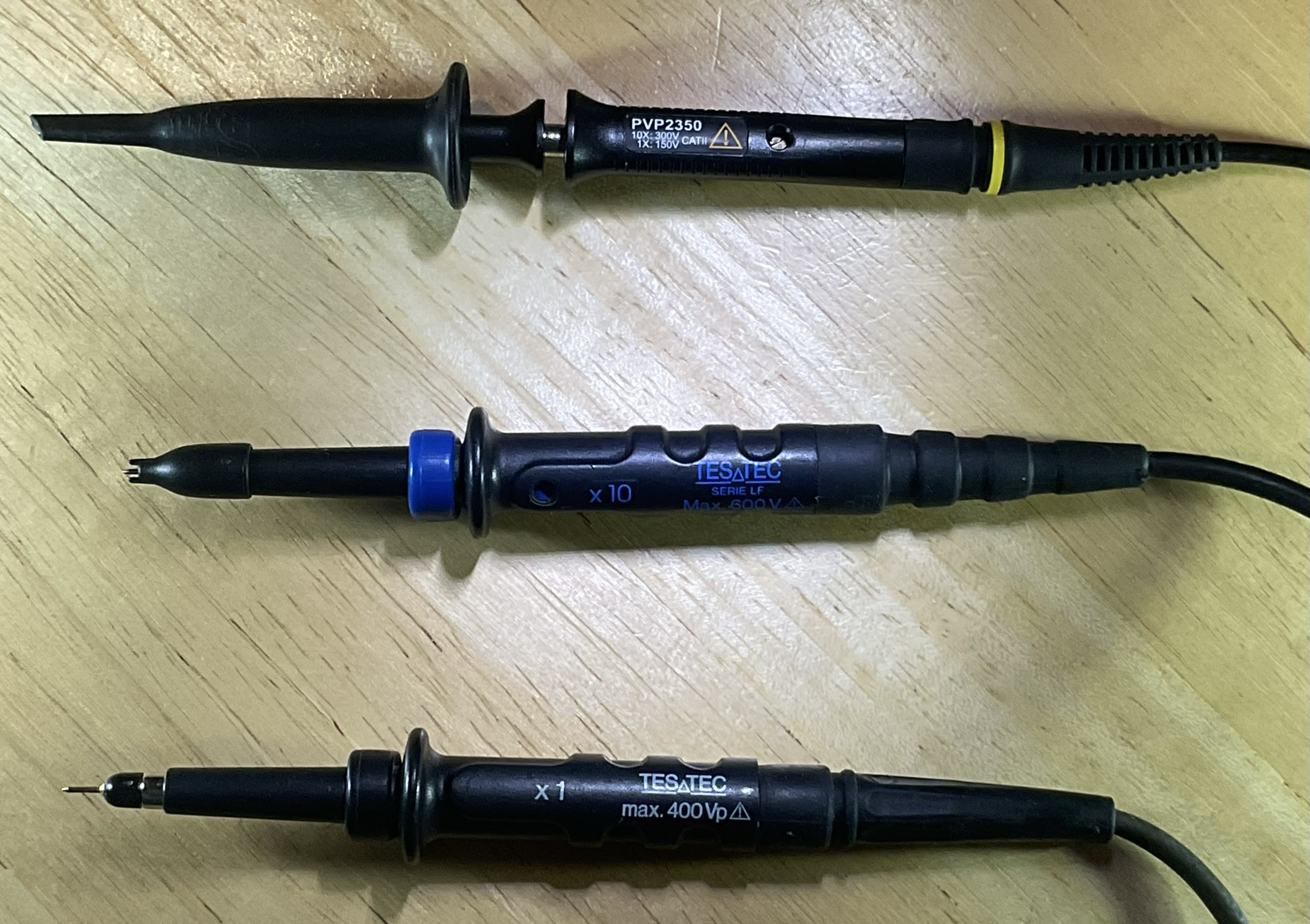 Dieser Alt-Text wurde noch nicht überprüft.
<ol>
<li>
<p>Kurzzusammenfassung: Drei schwarze Oszilloskop‑Tastköpfe liegen parallel auf einer hellen Holzoberfläche.</p>
</li>
<li>
<p>Detailbeschreibung: Die drei Sonden sind horizontal ausgerichtet, mit den Spitzen nach links und den Kabeln nach rechts. Oben befindet sich ein schwarzer Tastkopf mit der Aufschrift „PVP2350“, Warnsymbol und gelbem Ring an der Zugentlastung; die Spitze ist mit einer länglichen schwarzen Schutzkappe versehen. In der Mitte liegt ein schwarzer Tastkopf mit blauem Einstellring und der blauen Beschriftung „x10“, „TESATEC“, „SERIE LF“, „Max. 600V“; an der Spitze ist eine kleine Haken- oder Steckspitze sichtbar, teils von einer kurzen Kappe umgeben. Unten liegt ein weiterer schwarzer Tastkopf mit weiß/blauer Beschriftung „x1“, „TESATEC“, „max. 400Vp“; hier ist eine feine, nadelförmige Metallspitze freiliegt. Die Gehäuse haben gerippte Griffflächen, die Kabel führen nach rechts aus dem Bild, und die Holzoberfläche zeigt deutliche Maserung."></p>
 <figcaption>Abbildung A-2.3.4: Tastköpfe mit verschiedenen Prüfspitzen. Die Krokodilklemmen wurden für diese Aufnahme abgenommen.</figcaption>
</li>
</ol>
</figure>
</aside>
Die einfachsten Tastköpfe verbinden die Prüfspitze direkt mit dem Messeingang. Man spricht von 1:1-Tastköpfen, weil die an der Spitze anliegende Spannung unverändert zum Oszilloskop gelangt. Tastköpfe für hohe Frequenzen sind aufwändiger gebaut. Sie teilen die Eingangsspannung auf einen kleineren Wert, oft ein Zehntel, herunter. Wenn man mit so einem 10:1-Tastkopf eine Spannung von 10 Volt misst, wird auf dem Bildschirm 1 Volt angezeigt.
<aside class=