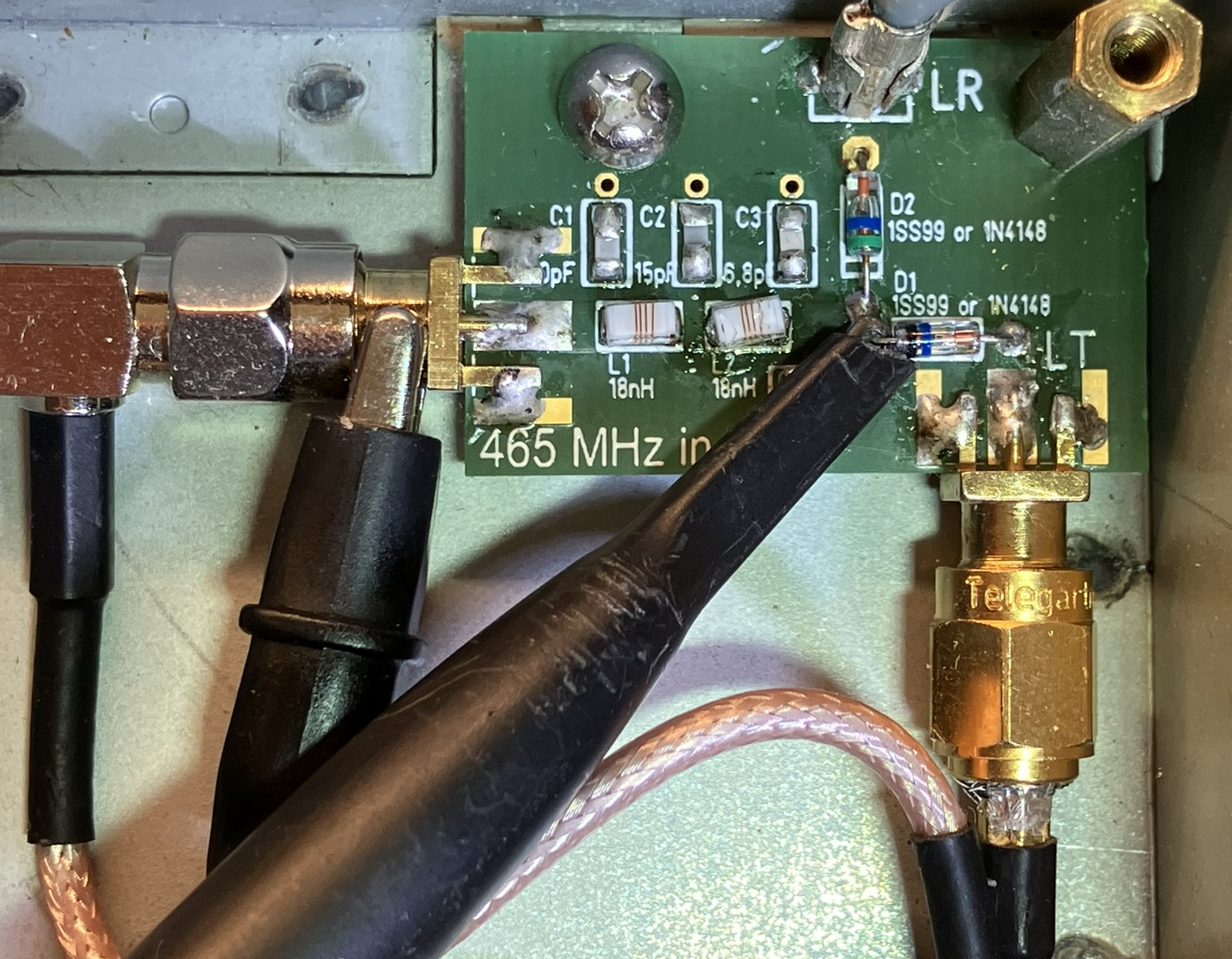 Dieser Alt-Text wurde noch nicht überprüft.
<ol>
<li>
<p>Kurzfassung: Geöffnete Elektronikbaugruppe in einem Metallgehäuse mit einer kleinen grünen Leiterplatte, mehreren Koax‑Steckverbindern und angesetzten Messleitungen.</p>
</li>
<li>
<p>Detaillierte Beschreibung: In der rechten Bildhälfte liegt eine grüne Leiterplatte mit weißer Beschriftung; sichtbar sind u. a. Bezeichnungen C1, C2, C3, L1, L2, D1, D2 sowie der Aufdruck „465 MHz in“. Neben den Markierungen für D1/D2 sind zwei kleine bedrahtete Bauteile mit Farbringen eingelötet; daneben liegen weitere bestückte und unbestückte SMD‑Plätze. Am rechten Rand ragt ein goldfarbener, verschraubter Koax‑Steckverbinder (SMA‑Bauform) nach unten aus dem Gehäuse; auf dem Sechskant steht „Telegärtner“. Links führt ein schwarzes Koaxkabel über einen rechtwinkligen metallischen Adapter auf die Platine. Über der Platine sind zwei schwarze Tastköpfe oder Leitungen angesetzt; ein spitzer schwarzer Prüfstift berührt einen Lötpunkt neben den beschrifteten Bauteilen. Die Platine ist mit mehreren Lötstellen, etwas Flussmittelrückständen und einer Schraube am Gehäuse befestigt; der Hintergrund besteht aus blankem Metall. Ein rosafarben ummanteltes Koaxkabel verläuft von unten rechts zum goldfarbenen Stecker."></p>
 <figcaption>Abbildung A-2.3.3: Messung mit einem Tastkopf. Zwischen den Dioden D1 und D2 ist die Prüfspitze zu sehen und weiter links die Krokodilklemme für die Masseverbindung.</figcaption>
</li>
</ol>
</figure>
</aside>
<aside class=