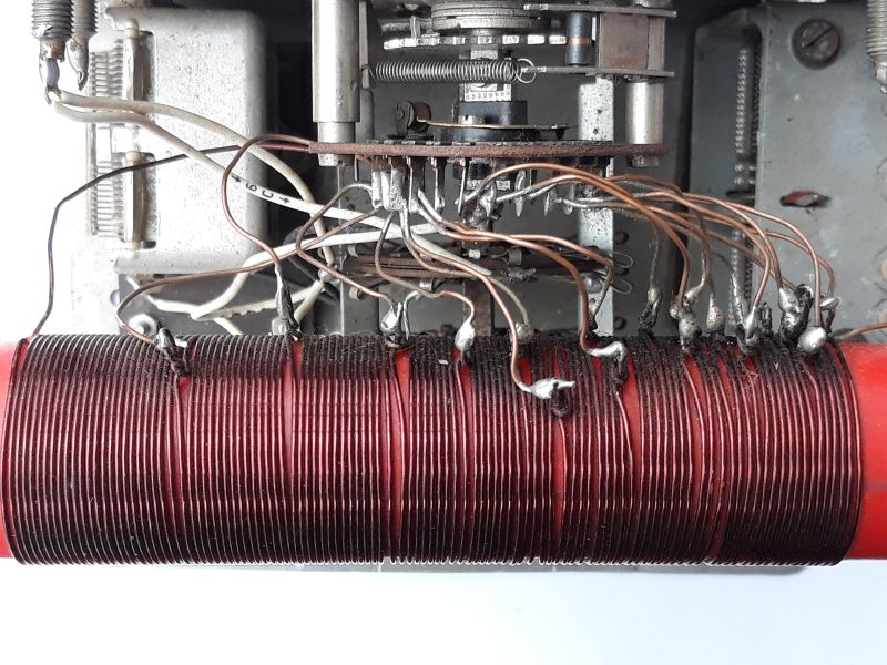 Dieser Alt-Text wurde noch nicht überprüft.
<p>Zusammenfassung: Nahaufnahme des Innenraums eines elektronischen Geräts mit einer dicht gewickelten roten Zylinderspule und zahlreichen Drahtabgriffen, die zu einem mehrstufigen Drehschalter im Metallchassis führen.</p>
<p>Detaillierte Beschreibung: Im unteren Bildbereich liegt horizontal eine rote, zylindrische Spule, eng mit dunkelrotem Kupferdraht bewickelt. Entlang der oberen Spulenseite sind viele Abgriffe sichtbar: dünne, bräunliche lackierte Drähte, die an einzelnen Windungen angelötet sind; an den Lötstellen sitzen silbrig glänzende Lottropfen, teils mit dunklen Verfärbungen an der Umgebung. Die Abgriffe verlaufen bogenförmig nach oben und vorne zu einem gestapelten, kreisförmigen Drehschalter mit mehreren Isolatorscheiben und radial angeordneten Lötfahnen. In der Schaltermitte sitzt eine Metallwelle mit Federn und kleinen mechanischen Teilen. Links und rechts erkennt man das graue Metallchassis mit Schrauben, Federn und Halterungen; einzelne weiße und graue Leitungen führen in den Aufbau. Der Hintergrund ist überwiegend grau-metallisch, unter der Spule ist ein heller, weißer Untergrund zu sehen. Der Gesamteindruck ist technisch, gebraucht und funktional, mit leicht unordentlich geführten Drähten.">
<figcaption>Abbildung NEA-9.8.1: Spule mit 14 Anzapfungen in einem selbstgebauten Antennenanpassgerät</figcaption></p>
</figure>
Die Teilabschnitte zwischen den Anzapfungen können wie einzelne Spulen gesehen werden, deren Induktivität, durch Umschaltung der Anzapfungen,  in Schritten erhöht werden kann. Die Reihenschaltung aller Teilabschnitte ergibt dann die Induktivität der Gesamtspule.
</aside>
Achte bei der folgenden Frage darauf alle Werte der Berechnung in die gleiche Einheit umzurechnen!
<section class=