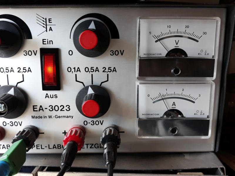 Dieser Alt-Text wurde noch nicht überprüft.
<ol>
<li>
<p>Zusammenfassung: Nahaufnahme der Frontplatte eines Geräts EA‑3023 mit zwei analogen Anzeigen (Volt und Ampere), zwei großen Drehknöpfen, einem rot leuchtenden Wippschalter und mehreren farbigen Anschlussbuchsen mit eingesteckten Leitungen.</p>
</li>
<li>
<p>Detaillierte Beschreibung: Links sind zwei schwarze Drehknöpfe mit roten Mitten zu sehen; um den oberen Knopf ist „0 … 30V“ markiert, um den unteren „0,1A 0,5A 2,5A“. Dazwischen befindet sich ein rechteckiger, rot beleuchteter Wippschalter mit der Beschriftung „Ein“ oben und „Aus“ unten. Darüber steht ein stilisiertes „EA“-Logo, daneben mehrfach „30V“. Im unteren linken Bereich steht „EA‑3023“ und „Made in W.-Germany“. Entlang der unteren Kante sind mehrere 4‑mm‑Buchsen und Stecker sichtbar: u. a. ein grüner, ein roter und ein schwarzer Stecker mit angeschlossenen Kabeln; neben den Buchsen ist „0‑30V“ aufgedruckt. Rechts sind zwei übereinander angeordnete analoge Messwerke der Marke „MONACOR“: oben mit der Skala „0–30“ und dem Symbol „V“ (Volt), unten mit der Skala „0–3“ und dem Symbol „A“ (Ampere); beide Instrumente tragen die Kennzeichnung „Kl. 2.0“. Das Gehäuse wirkt metallisch/silbern, die Beschriftungen sind schwarz, Bedienelemente überwiegend schwarz mit roten Akzenten."></p>
 <figcaption>Abbildung NEA-10.2.2: Labornetzgerät mit eingestellter Strombegrenzung auf $500&nbsp;mA$</figcaption>
</li>
</ol>
</figure>
<p>In Labornetzgeräten ist eine Strombegrenzung eingebaut, d.h. übersteigt der Laststrom eine maximale Stromstärke, wird die Klemmenspannung so abgesenkt, dass der Laststrom konstant bleibt. Dies entspricht der Funktion einer Konstantstromquelle -- Bei Kurzschluss an den Ausgangsklemmen fließt der eingestellte maximale Strom.</p>
</aside>
Eine ideale Konstantstromquelle liefert unabhängig von der angeschlossenen Last einen konstanten Dauerstrom. In der Theorie ist dies bei einem unendlichen Innenwiderstand möglich. In der Praxis haben Stromquellen einen sehr hohen Innenwiderstand.
<aside class=