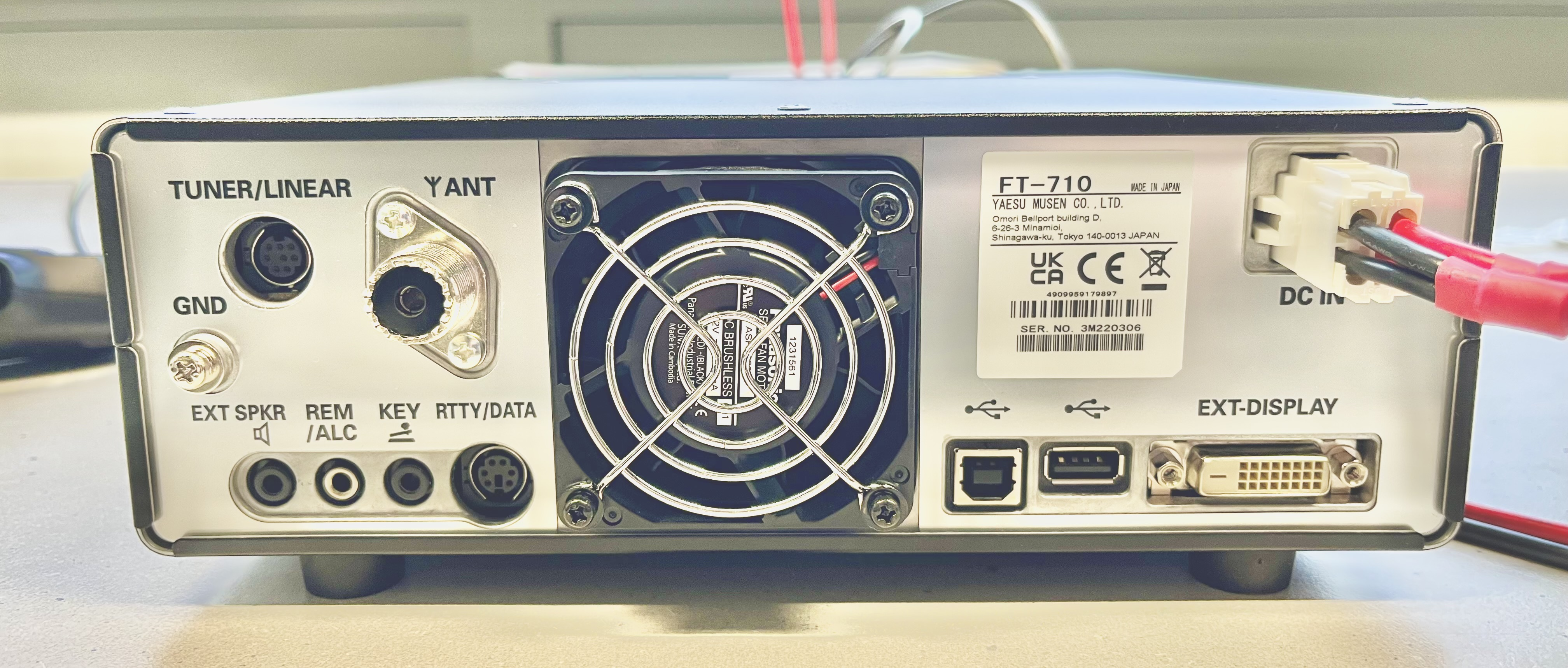Dieser Alt-Text wurde noch nicht überprüft.
<ol>
<li>
<p>Kurze Zusammenfassung: Rückseite eines elektronischen Geräts (Modell FT‑710) mit vielen Anschlüssen, zentralem Lüftergitter und angeschlossenem DC‑Stromkabel.</p>
</li>
<li>
<p>Detaillierte Beschreibung: Das Gerät steht auf einem hellen Tisch; im Zentrum befindet sich ein großer, runder, mit vier Schrauben befestigter Lüfter mit Metall-Schutzgitter und sichtbarem „Brushless Fan“-Aufkleber. Links sind mehrere Buchsen und Beschriftungen: oben „TUNER/LINEAR“ an einer runden mehrpoligen Mini‑DIN‑Buchse, darunter „GND“ an einem Massebolzen mit Rändelmutter, daneben „Y ANT“ bei einer großen koaxialen SO‑239‑Buchse. In der unteren linken Reihe stehen nebeneinander Klinkenbuchsen und Mini‑DIN‑Buchsen, beschriftet mit „EXT SPKR“, „REM/ALC“, „KEY“ und „RTTY/DATA“. Rechts neben dem Lüfter klebt ein weißes Typenschild mit der Aufschrift „FT‑710“, Hersteller‑ und Zulassungszeichen sowie einer Seriennummer. Ganz rechts oben ist „DC IN“ mit einem weißen Steckverbinder zu sehen, in den rote und schwarze Stromkabel eingesteckt sind. Unten rechts ist der Bereich „EXT‑DISPLAY“ mit mehreren Schnittstellen: zwei USB‑Anschlüsse (links ein quadratischer Typ‑B, daneben ein rechteckiger Typ‑A), ein kleiner gewindeter Koax‑Anschluss und ein weißer DVI‑Anschluss mit zwei Befestigungsschrauben. Der Hintergrund ist unscharf und zeigt eine helle Umgebung."></p>
 <figcaption>Abbildung NE-16.7.1: Funkgerät mit DATA-Port</figcaption>
</li>
</ol>
</figure>
<figure class=