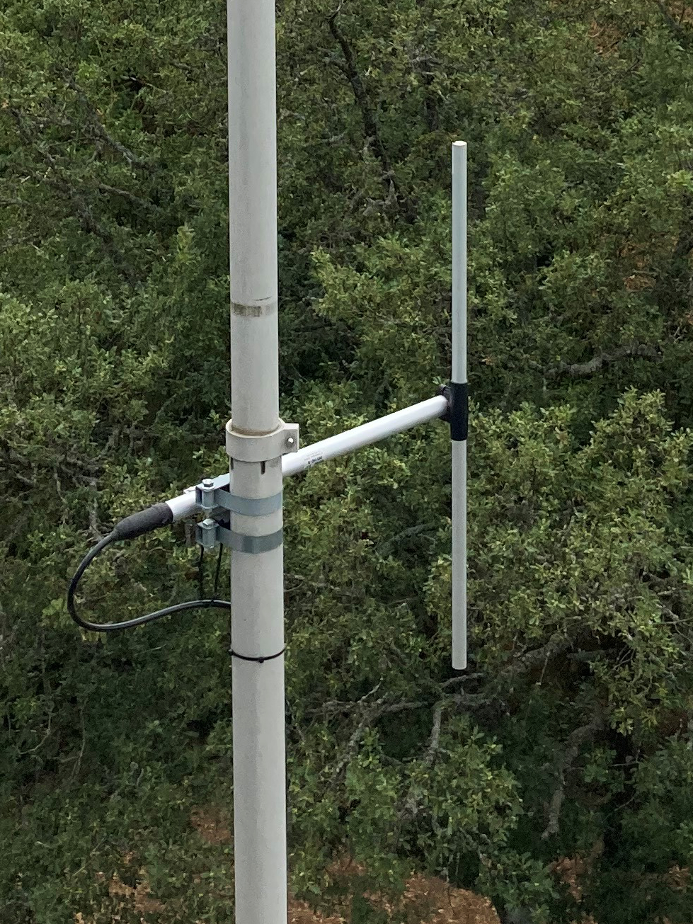 Dieser Alt-Text wurde noch nicht überprüft.
<ol>
<li>
<p>Kurzzusammenfassung: An einem hellen Mast ist eine kleine Antenne mit zwei parallelen vertikalen Stäben montiert, angeschlossen über ein schwarzes Koaxkabel; im Hintergrund dichter Baumbestand.</p>
</li>
<li>
<p>Detaillierte Beschreibung: Ein senkrechter, hellgrauer Mast verläuft links im Bild und zeigt leichte Gebrauchsspuren. Mit zwei Metallbandschellen ist daran ein kurzer, waagerechter Ausleger befestigt. Am freien Ende des Auslegers stehen zwei schlanke, helle Stäbe parallel nach oben; der hintere Stab ist länger, der vordere etwas kürzer und mit geringem Abstand davor. Am Übergang zwischen Mast und Ausleger sitzt eine kleine Halterung, in die ein schwarzes Koaxialkabel mit Stecker eingeführt ist; die Anschlusspunkte sind mit dunklem Band oder Schrumpfschlauch abgedichtet. Das Kabel führt in einer Schlaufe nach unten. Der Hintergrund besteht vollständig aus dichtem, grünem Laub von Bäumen; nahe dem unteren Bildrand ist etwas brauner Boden durch das Geäst zu erkennen."></p>
 <figcaption>Abbildung NEA-19.9.5: Vertikaler $\frac{\lambda}{2}$-Dipol</figcaption>
</li>
</ol>
</figure>
</aside>
---
<p>Einen Spezialfall einer Vertikalantenne stellt die gegen Erde (oder einer Fahrzeugkarosserie) erregte $5/8 \lambda$-Antenne dar (vgl. Abbildung <a class=