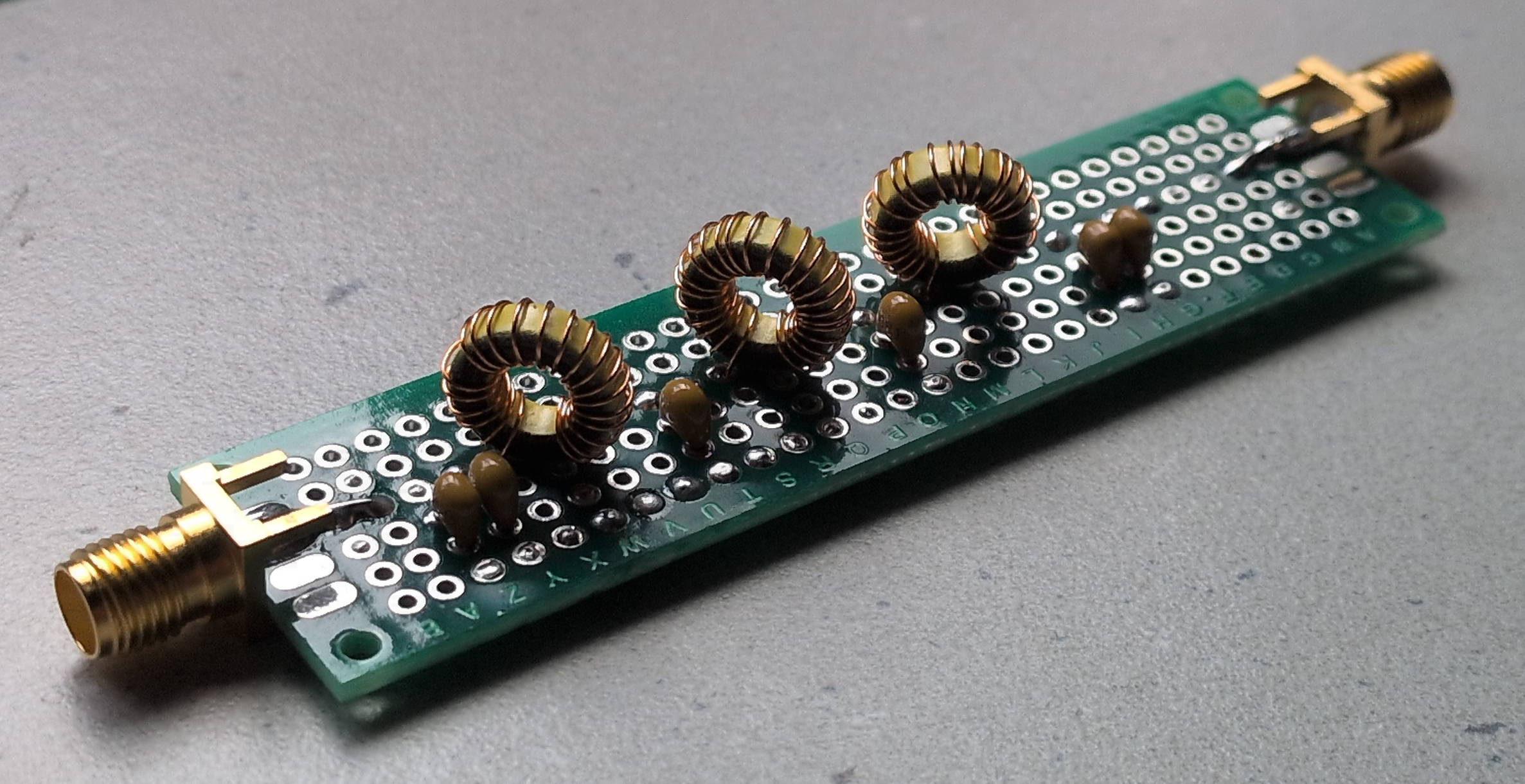 Dieser Alt-Text wurde noch nicht überprüft.
<ol>
<li>
<p>Zusammenfassung: Langer, schmaler Elektronikaufbau auf grünem Lochrasterboard mit drei ringförmigen Kupferspulen und mehreren kleinen braunen Bauteilen zwischen zwei goldfarbenen Koaxanschlüssen.</p>
</li>
<li>
<p>Detaillierte Beschreibung: Auf einem rechteckigen, grünen Lochrasterboard mit Reihen aus runden Lötaugen sind drei gleichartige, ringförmige Spulen aus lackiertem Kupferdraht auf gelblichen Kernen in einer Linie angeordnet. Dazwischen und daneben sitzen mehrere kleine, braun glänzende, tropfenförmige Bauteile (per Drahtanschluss eingelötet). An beiden Stirnseiten des Boards befinden sich goldfarbene, koaxiale Buchsen/Stecker mit verlöteten Mittelkontakten und seitlichen Lötfahnen zur Masse. Die Lötstellen sind sichtbar, teils mit dunklen Flussmittelspuren. Das Board liegt schräg auf einer grauen, leicht strukturierten Oberfläche; die Beleuchtung kommt von oben links und erzeugt weiche Schatten."></p>
 <figcaption>Abbildung NE-11.1.10: Selbstgebautes Tiefpassfilter</figcaption>
</li>
</ol>
</figure>
Die oben genannten Filter lassen sich natürlich wunderbar für alle Frequenzbereiche selbst berechnen und bauen. In der Formelsammlung finden sich die benötigten Formeln, wobei es natürlich auch viele Bauvorschläge und Berechnungsprogramme gibt. Die benötigten Spulen lassen sich dazu oft leicht selbst herstellen. Hierfür reicht bei kleinen Spulenwerten ein kleiner Vorrat an $0,8&nbsp;mm$-Kupferlackdraht für stabile Luftspulen aus. Bei großen Spulenwerten z.B. für die Kurzwellenbereiche kommt man mit $0,2&nbsp;mm$-Kupferlackdraht und Kernmaterial mit entsprechenden $A_\text{L}$-Werten aus, um sich jederzeit die richtigen Werte selbst herstellen zu können. Die benötigten Größen, Wicklungen usw. bekommt man über Formelsammlung, Bauvorschläge oder Berechnungsprogramme ebenfalls meist leicht heraus.
</aside>
---
<p>Nun haben wir einfache RC- und LC-Glieder als Hoch- und Tiefpassfilter kennengelernt. Aus Kondensatoren und Spulen lassen sich jedoch noch weitere Filtertypen realisieren, die über reine Hoch- und Tiefpässe hinausgehen. Diese wollen wir uns nun im zweiten Teil genauer ansehen, nämlich die sogenannten <em>Schwingkreise</em>.</p>
<aside class=